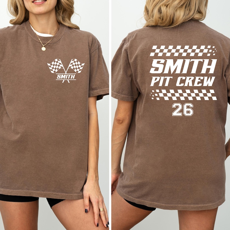 以下が含まれることがあります： レーシングテーマの茶色のTシャツ。前面には、交差したチェッカーフラッグと「SMITH」の文字が描かれています。背面には「SMITH PIT CREW」と数字の「26」、チェッカーフラッグのデザインが表示されています。