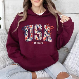 May include: Burgundy hooded sweatshirt with a floral print of the word "USA" and the text "EST. 1776" below it.