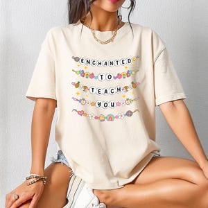 May include: A beige t-shirt with the words "ENCHANTED TO TEACH YOU" in a colorful, whimsical font. The text is arranged in a stacked format, with each word on a separate line. Decorative elements like flowers, fruits, and rainbows surround the text.