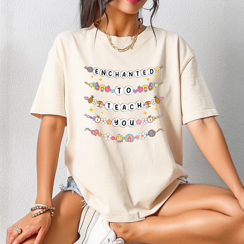 May include: A beige t-shirt with the words "ENCHANTED TO TEACH YOU" in a colorful, whimsical font. The text is arranged in a stacked format, with each word on a separate line. Decorative elements like flowers, fruits, and rainbows surround the text.