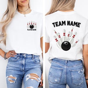 May include: White t-shirt featuring a bowling design. The front displays a bowling ball and pins with "PLAYER NAME" text. The back shows a bowling ball and pins with "TEAM NAME" text. The shirt is paired with blue jeans.