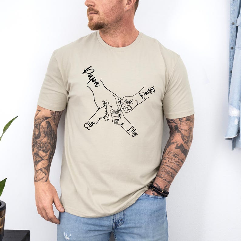 May include: A beige t-shirt with a black line drawing of four hands holding each other. The names "Papa", "Daisy", "Ella", and "Lily" are written on the hands.