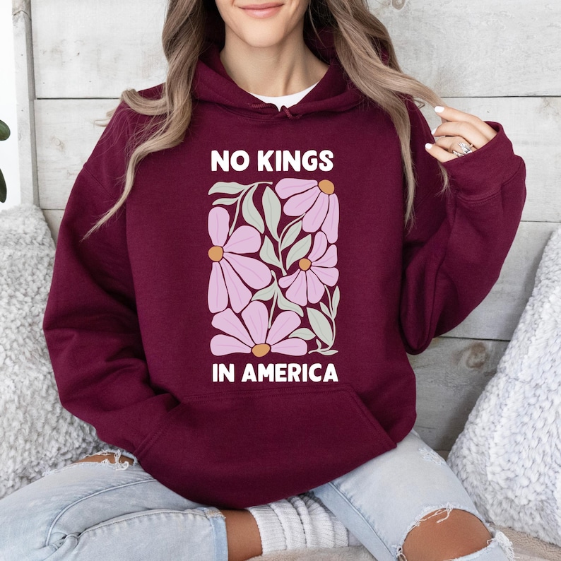 May include: Burgundy hoodie with a floral design and the text "NO KINGS IN AMERICA" in white. The graphic features pink flowers and green leaves. The hoodie has a front pocket and a hood.