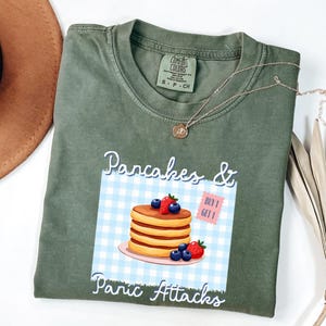 Peut inclure: T-shirt vert olive avec l'inscription "Pancakes & Panic Attacks" et un dessin de crêpes avec des myrtilles et des fraises. Le t-shirt a un col rond et des manches courtes. Un collier doré est posé sur le t-shirt.