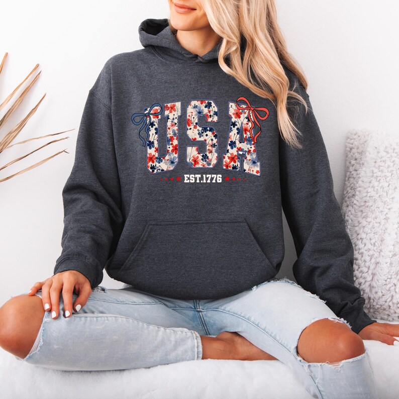 May include: A dark heather grey hoodie with a floral print of the word "USA" and the text "EST. 1776".