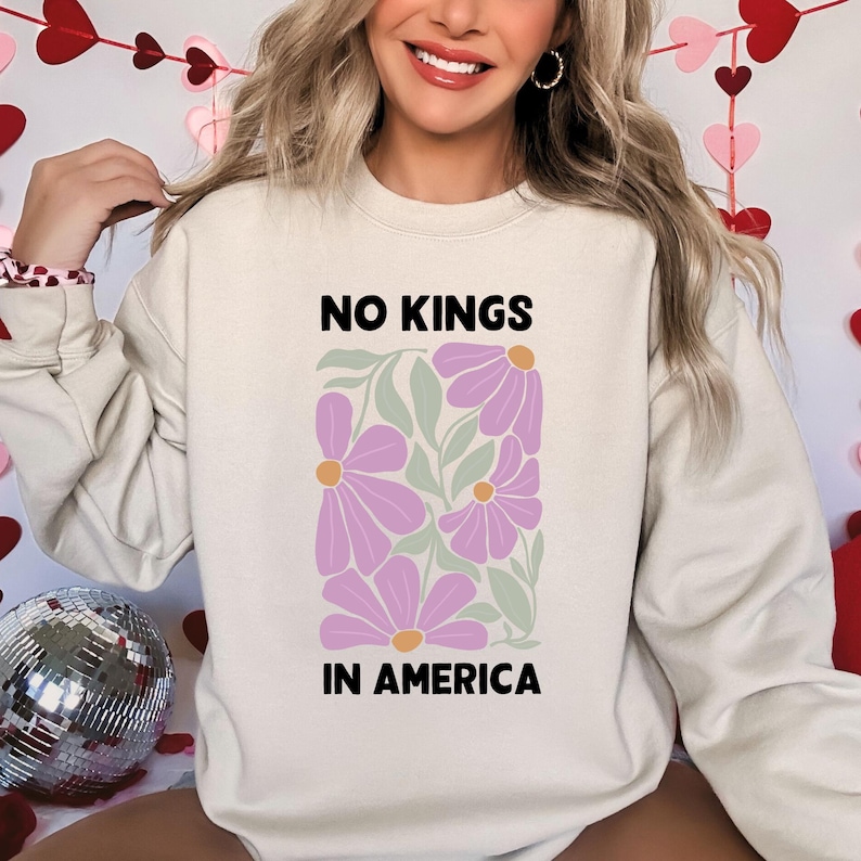 May include: A cream-coloured sweatshirt featuring the text "NO KINGS IN AMERICA" above a floral design of purple flowers and green leaves. The sweatshirt is a casual, comfortable style.