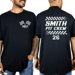 以下が含まれることがあります： 白のグラフィックが施された黒いTシャツ。前面には2つのチェッカーフラッグと「SMITH」の文字が描かれています。背面には「SMITH PIT CREW」と数字の「26」がチェッカーフラッグのアクセントとともに表示されています。カジュアルなクルーネックTシャツ。
