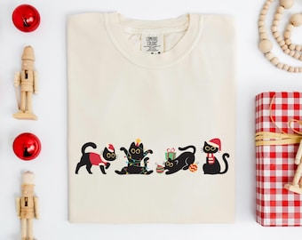 Puede incluir: Camiseta de color crema con un diseño festivo de gatos negros con atuendo navideño. Los gatos están decorados con luces navideñas, gorros de Papá Noel y regalos. La camiseta está rodeada de adornos navideños y un regalo envuelto.