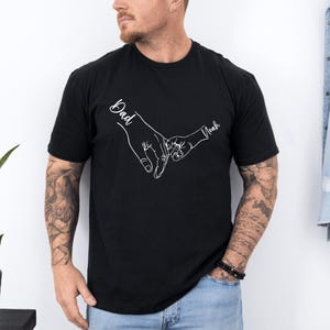May include: Black t-shirt with a white outline of two hands holding each other. The text "Dad" is above one hand and "Noah" is above the other.