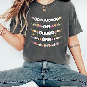 May include: Dark gray t-shirt with the words "ENCHANTED TO TEACH YOU" in white letters. The text is decorated with colorful illustrations of hearts, stars, flowers, and rainbows. The shirt is a comfortable, casual style.