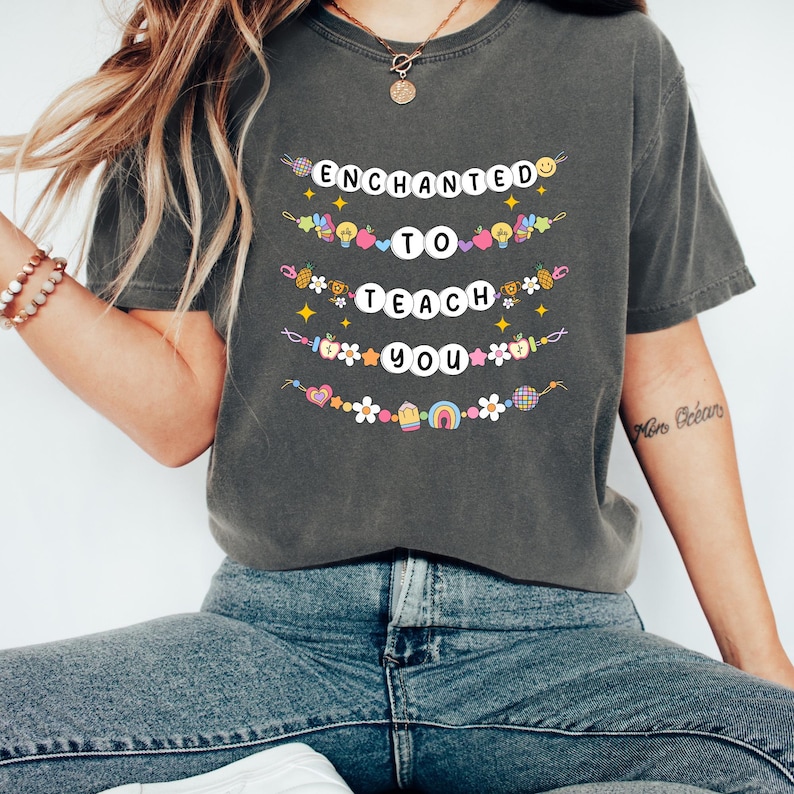 May include: Dark gray t-shirt with the words "ENCHANTED TO TEACH YOU" in white letters. The text is decorated with colorful illustrations of hearts, stars, flowers, and rainbows. The shirt is a comfortable, casual style.