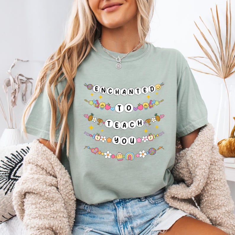 May include: A light green t-shirt with the words "ENCHANTED TO TEACH YOU" in a whimsical, colorful design. The design includes various illustrations like fruits, flowers, and rainbows. The shirt is a casual, everyday wear item.