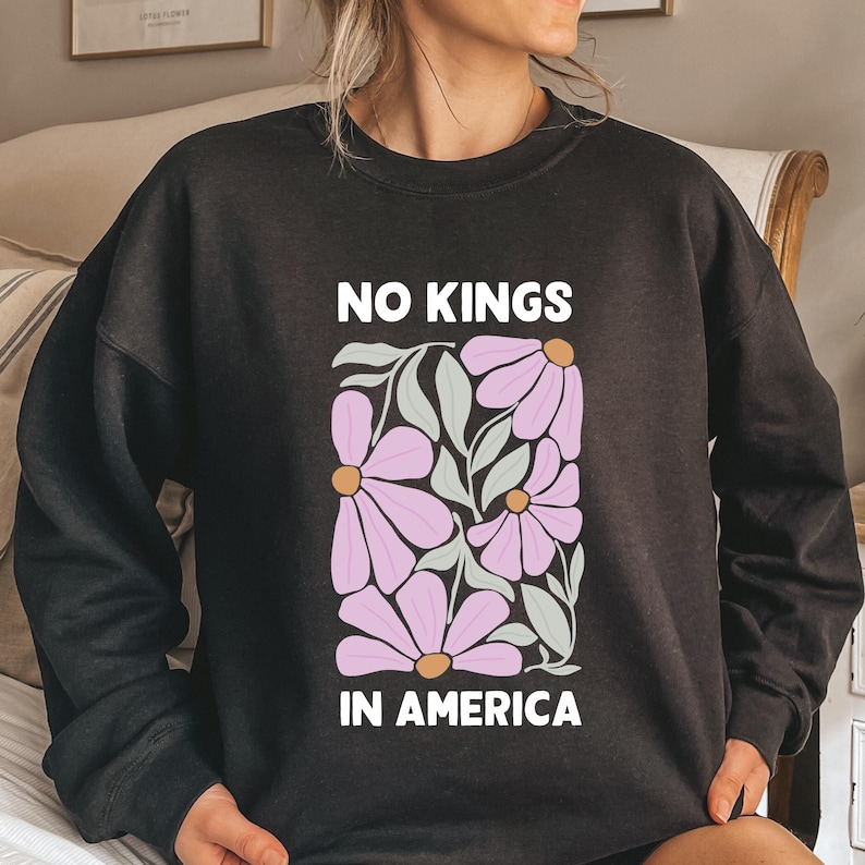 May include: Dark grey sweatshirt with the slogan "NO KINGS IN AMERICA" in white. The design includes a rectangular floral pattern with pink flowers, green leaves, and yellow centres. A relaxed fit top.