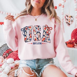 May include: Pink sweatshirt with a floral print of the word "USA" and the text "EST. 1776" below it.