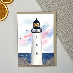 Può includere: Dipinto ad acquerello di un faro bianco con la parte superiore nera, ambientato su un cielo blu e rosa. Il faro è su una carta con bordo marrone, con piccoli uccelli neri che volano sopra. Il mare è blu, su sfondo neutro.
