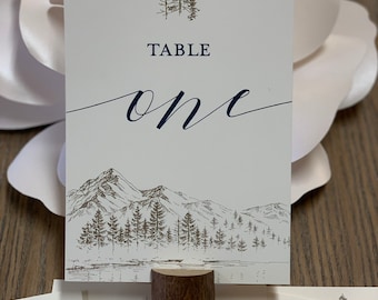 May include: A white card with a brown mountain scene and a dark blue cursive font that says "TABLE one". The card is held up by a brown wooden stand.