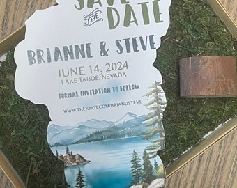 Puede incluir: Tarjeta de guardar la fecha con una ilustración de acuarela del lago Tahoe, montañas y pinos. La tarjeta tiene forma de lago y tiene el texto "Save the Date" en verde y "Brian & Steve" en plata. La fecha es el 14 de junio de 2024 y la ubicación es el lago Tahoe, Nevada. El texto "Formal Invitation to Follow" está incluido, junto con el sitio web www.theknot.com/briandsteve.