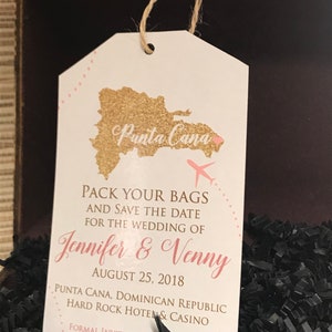 May include: A white luggage tag with a gold glitter outline of the Dominican Republic and the text "Punta Cana". The tag also includes a pink heart, a small airplane, and the text "Pack your bags and save the date for the wedding of Jennifer & Venny August 25, 2018 Punta Cana, Dominican Republic Hard Rock Hotel & Casino Formal Invitation To".