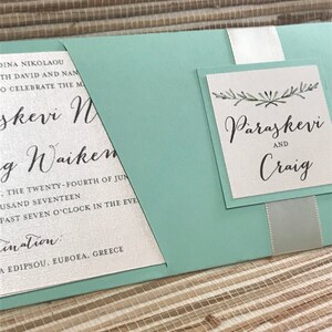 May include: A light blue wedding invitation with white ribbon and a white card with the text "Paraskevi and Craig" in black cursive font.
