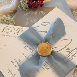 May include: A white wedding invitation with a blue ribbon and a gold wax seal. The invitation says "June 16, 2022" and "three o'clock" in gray text. The invitation also includes the text "reception to follow".