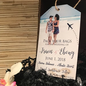 May include: A luggage tag with a blue background and a photo of a couple in front of a beach. The text on the tag reads "Pack Your Bags for the Wedding of Travis & Ebony June 1, 2018 Nassau Bahamas - Atlantis Resort Formal Invitation to Follow COM/THEWNBREA VOW2018".