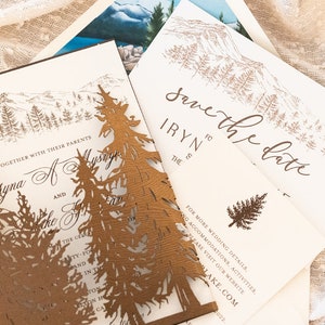 May include: Save the date wedding invitation with a mountain and forest design. The invitation is printed on white cardstock with brown ink. The text reads "Save the Date" and "IRYN to THE".