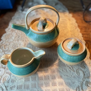 May include: A ceramic tea set with a teapot, creamer, and sugar bowl. The set features a teal and tan color scheme with a natural handle and lid. The teapot has a spout and a curved handle. The sugar bowl has a lid with a small handle.