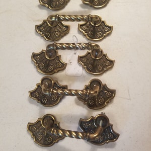 May include: Five antique brass drawer pulls. Each pull features a decorative floral design on the ends and a twisted handle. The pulls are a warm, aged brass color, suggesting vintage or retro style. These are suitable for furniture restoration or adding a vintage touch.
