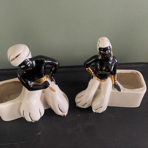 May include: Two ceramic figurines of figures with black skin, white pants, and white headwear. The figures are posed in front of rectangular planters. The figures have gold accents on their clothing.