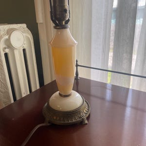 May include: A vintage table lamp with a white and yellow glass body and a decorative bronze base. The lamp has a classic design with a bulb at the top and a power cord. The lamp is on a dark wooden surface.