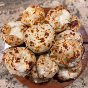 May include: A wooden plate holds a pile of baked cheese bread. The bread is golden brown with darker, toasted spots on top. The interior of the bread appears soft and filled with melted cheese, creating a delicious and appetizing presentation.