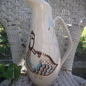 May include: A tall, off-white ceramic pitcher with a speckled finish and a curved handle. The body features a painted brown and blue bird illustration with a stylized wing pattern and a small blue dandelion seed puff floating above the bird.