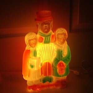 May include: A glowing, molded plastic light fixture depicting three figures singing together. The central figure wears a hat and plays an accordion, while the two flanking figures hold songbooks. The piece emits a warm orange light.