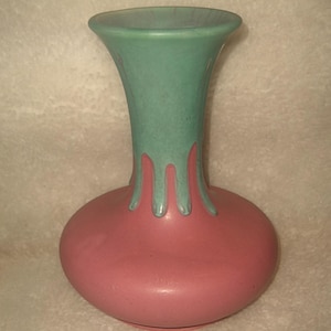 May include: A ceramic vase with a round base and a flared neck. The vase is primarily a dusty rose color, with a turquoise-green neck and drips of the same color cascading down the body. The vase has a smooth, matte finish.