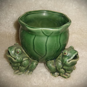 May include: A green ceramic planter featuring a central bowl with a cabbage leaf texture and a glossy crackle finish. Two sculpted green frogs with detailed skin textures and open mouths are positioned on opposite sides of the base.