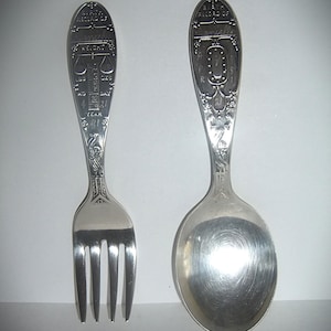 May include: A silver-toned metal fork and spoon set featuring engraved birth record details. The fork handle displays weight in lbs and oz, while the spoon handle features a clock face. Both pieces include the name Danielle and the year 81.