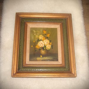 May include: A framed floral still life painting. The artwork features a bouquet of yellow and cream roses in a gold vase. The painting is set within a gold and green decorative frame.