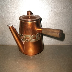 May include: A copper-colored teapot with a wooden handle and a decorative silver-colored design. The teapot has a spout and a lid with a small knob. The handle is made of light-colored wood.