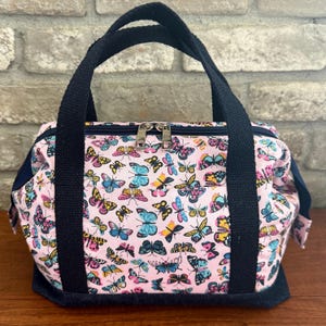 May include: A small, rectangular handbag with a pink background covered in colorful butterfly patterns. The bag has a navy blue base and handles, with a silver zipper closure. The bag is sitting on a wooden surface.