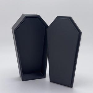 May include: A black, open coffin-shaped box with a textured surface. The lid is separate from the base, revealing the interior. The coffin is made of a solid, dark material, and is set against a plain background.