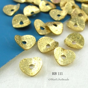 May include: A close-up of a pile of gold-colored heart-shaped metal beads with a hole in the center. The beads have a textured surface and are scattered on a blue background. The text "RB 111" and "©BlueEchoBeads" is visible in the lower right corner of the image.