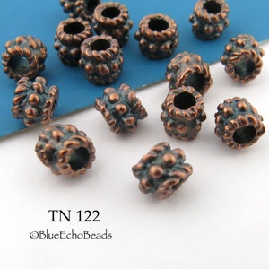 May include: A close-up of a group of copper-colored metal beads with a textured surface. The beads have a hole in the center and are arranged on a blue and white surface. The text "TN 122 ©BlueEchoBeads" is visible in the lower part of the image.