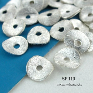 May include: A close-up of a pile of silver-colored metal beads with a textured surface. The beads are shaped like a triangle with a round hole in the center. The beads are on a blue surface. SP110 ©BlueEchoBeads