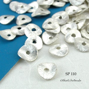May include: A pile of silver-colored metal beads with a textured surface and a hole in the center. The beads are shaped like a triangle with rounded edges. The beads are scattered on a blue and white surface. SP 110 ©BlueEchoBeads
