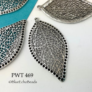 May include: Silver filigree leaf pendant with a detailed design. The pendant has a rounded edge and a hole at the top for hanging.