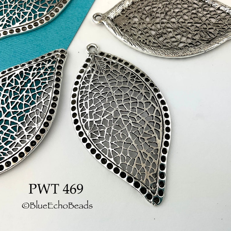 May include: Silver filigree leaf pendant with a detailed design. The pendant has a rounded edge and a hole at the top for hanging.