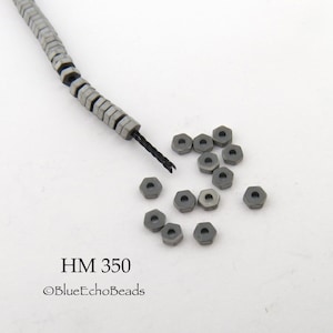 May include: A close-up of a pile of small, gray, metal hexagonal beads with a hole in the center. The beads are scattered on a white surface. A strand of the same beads is visible in the background. The text "HM 350 ©BlueEchoBeads" is visible in the lower left corner of the image.