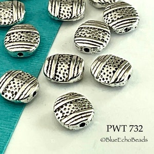May include: Silver oval-shaped beads with a textured surface and a raised design. The beads have a hole in the center for stringing. PWT 732 ©BlueEchoBeads