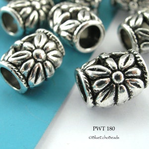 May include: Close-up of several silver-colored metal beads. Each bead has a cylindrical shape with a floral design around the middle. The beads have a distressed, antique finish. The text "PWT 180" and "©BlueEchoBeads" are visible.