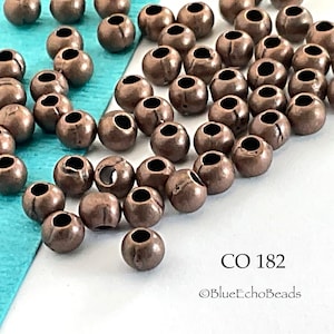 May include: A large quantity of small, round, copper-colored beads with a hole in the center. The beads are scattered on a white surface with a turquoise blue surface in the background. The text "CO 182" and "@BlueEchoBeads" is visible in the lower right corner of the image.
